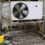 Exterior view of an industrial air conditioning unit with visible wear and rust, set against a shabby wall.